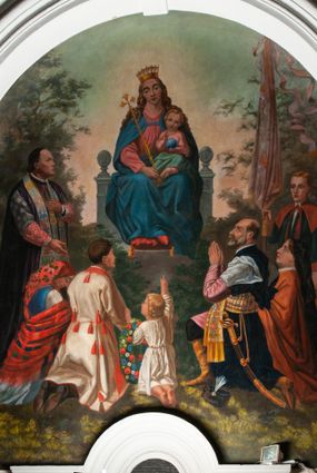 Zdjęcie nr 1: Fresk wkomponowany w arkadę zamkniętą półkoliście, w ścianie południowej prezbiterium, nad portalem do aneksu. Ukazuje siedzącą na tronie Matkę Boską z Dzieciątkiem na kolanach oraz klęczących i stojących przed nią od lewej proboszcza Józefa Krzemińskiego, rodzinę w stroju ludowym, Izabelę i Felicjana Szybalskich oraz chłopca z chorągwią w ręku. 