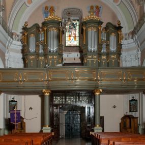 Zdjęcie nr 1: Prospekt organowy jest dwuczęściowy, czterowieżowy, zbudowany na wysokim cokole, o ośmiu rejestrach. Każda z części prospektu jest ujęta parą złoconych kolumn korynckich, stojących na ukośnie dostawionych postumentach z wolutowymi impostami. Kolumny podtrzymują spływy wolutowe zakończone wazonami płomienistymi (skrajne wieże), lub wybrzuszone łuki odcinkowe zakończone motywem rocaille&#039;m (wewnętrzne wieże). Cztery największe rejestry przysłonięte pozłacanymi kotarami. Kontuar wolnostojący, pomiędzy obiema częściami szafy organowej, na osi założenia.