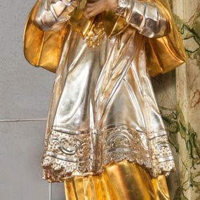 Zdjęcie nr 1: Rzeźba drążona i ścięta, ustawiona na podstawie na planie prostokąta. Święty przedstawiony został w całej postaci, frontalnie, z łukowato wygiętą sylwetką, w kontrapoście, z lewą nogą odciążoną. Obie ręce ma złożone na piersi. Głowę ma uniesioną i zwróconą delikatnie w lewo, twarz podłużną, o łagodnych rysach, wznosi uniesiony, nos wąski, usta grube, zamknięte, na policzkach zaznaczone rumieńce, broda silnie wyodrębniona, zaczerwieniona; głowa okolona krótkimi, kręconymi, brązowymi włosami. Jest ubrany w złotą albę, srebrną komżę dekorowaną u dołu pasem koronki oraz muncet, spod szat wystają czubki złotych butów. Na piersi ma płaskorzeźbione, gorejące serce. Alba i kapa kształtowane sztywno, komża drobnymi fałdami przylega do spodniego ubrania. Partie ciała są polichromowane naturalistyczne. 