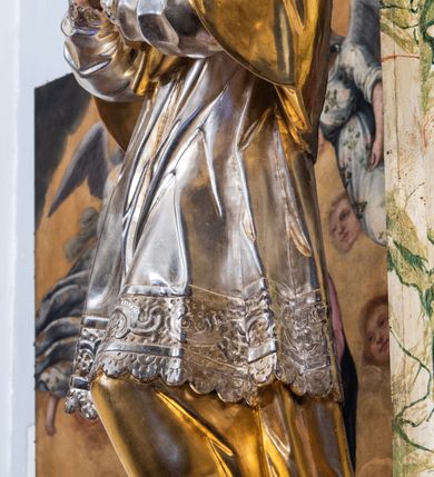 Zdjęcie nr 7: Rzeźba drążona i ścięta, ustawiona na podstawie na planie prostokąta. Święty przedstawiony został w całej postaci, frontalnie, z łukowato wygiętą sylwetką, w kontrapoście, z lewą nogą odciążoną. Obie ręce ma złożone na piersi. Głowę ma uniesioną i zwróconą delikatnie w lewo, twarz podłużną, o łagodnych rysach, wznosi uniesiony, nos wąski, usta grube, zamknięte, na policzkach zaznaczone rumieńce, broda silnie wyodrębniona, zaczerwieniona; głowa okolona krótkimi, kręconymi, brązowymi włosami. Jest ubrany w złotą albę, srebrną komżę dekorowaną u dołu pasem koronki oraz muncet, spod szat wystają czubki złotych butów. Na piersi ma płaskorzeźbione, gorejące serce. Alba i kapa kształtowane sztywno, komża drobnymi fałdami przylega do spodniego ubrania. Partie ciała są polichromowane naturalistyczne. 