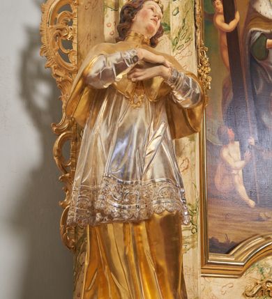 Zdjęcie nr 2: Rzeźba drążona i ścięta, ustawiona na podstawie na planie prostokąta. Święty przedstawiony został w całej postaci, frontalnie, z łukowato wygiętą sylwetką, w kontrapoście, z lewą nogą odciążoną. Obie ręce ma złożone na piersi. Głowę ma uniesioną i zwróconą delikatnie w lewo, twarz podłużną, o łagodnych rysach, wznosi uniesiony, nos wąski, usta grube, zamknięte, na policzkach zaznaczone rumieńce, broda silnie wyodrębniona, zaczerwieniona; głowa okolona krótkimi, kręconymi, brązowymi włosami. Jest ubrany w złotą albę, srebrną komżę dekorowaną u dołu pasem koronki oraz muncet, spod szat wystają czubki złotych butów. Na piersi ma płaskorzeźbione, gorejące serce. Alba i kapa kształtowane sztywno, komża drobnymi fałdami przylega do spodniego ubrania. Partie ciała są polichromowane naturalistyczne. 