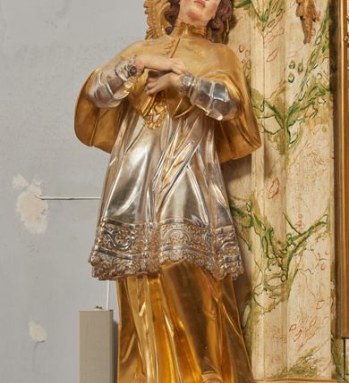 Zdjęcie nr 4: Rzeźba drążona i ścięta, ustawiona na podstawie na planie prostokąta. Święty przedstawiony został w całej postaci, frontalnie, z łukowato wygiętą sylwetką, w kontrapoście, z lewą nogą odciążoną. Obie ręce ma złożone na piersi. Głowę ma uniesioną i zwróconą delikatnie w lewo, twarz podłużną, o łagodnych rysach, wznosi uniesiony, nos wąski, usta grube, zamknięte, na policzkach zaznaczone rumieńce, broda silnie wyodrębniona, zaczerwieniona; głowa okolona krótkimi, kręconymi, brązowymi włosami. Jest ubrany w złotą albę, srebrną komżę dekorowaną u dołu pasem koronki oraz muncet, spod szat wystają czubki złotych butów. Na piersi ma płaskorzeźbione, gorejące serce. Alba i kapa kształtowane sztywno, komża drobnymi fałdami przylega do spodniego ubrania. Partie ciała są polichromowane naturalistyczne. 