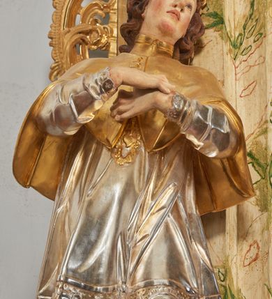 Zdjęcie nr 8: Rzeźba drążona i ścięta, ustawiona na podstawie na planie prostokąta. Święty przedstawiony został w całej postaci, frontalnie, z łukowato wygiętą sylwetką, w kontrapoście, z lewą nogą odciążoną. Obie ręce ma złożone na piersi. Głowę ma uniesioną i zwróconą delikatnie w lewo, twarz podłużną, o łagodnych rysach, wznosi uniesiony, nos wąski, usta grube, zamknięte, na policzkach zaznaczone rumieńce, broda silnie wyodrębniona, zaczerwieniona; głowa okolona krótkimi, kręconymi, brązowymi włosami. Jest ubrany w złotą albę, srebrną komżę dekorowaną u dołu pasem koronki oraz muncet, spod szat wystają czubki złotych butów. Na piersi ma płaskorzeźbione, gorejące serce. Alba i kapa kształtowane sztywno, komża drobnymi fałdami przylega do spodniego ubrania. Partie ciała są polichromowane naturalistyczne. 