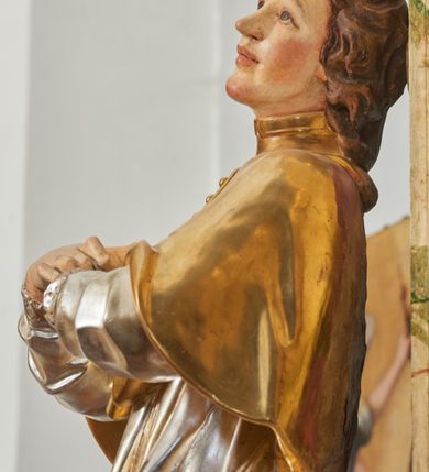 Zdjęcie nr 12: Rzeźba drążona i ścięta, ustawiona na podstawie na planie prostokąta. Święty przedstawiony został w całej postaci, frontalnie, z łukowato wygiętą sylwetką, w kontrapoście, z lewą nogą odciążoną. Obie ręce ma złożone na piersi. Głowę ma uniesioną i zwróconą delikatnie w lewo, twarz podłużną, o łagodnych rysach, wznosi uniesiony, nos wąski, usta grube, zamknięte, na policzkach zaznaczone rumieńce, broda silnie wyodrębniona, zaczerwieniona; głowa okolona krótkimi, kręconymi, brązowymi włosami. Jest ubrany w złotą albę, srebrną komżę dekorowaną u dołu pasem koronki oraz muncet, spod szat wystają czubki złotych butów. Na piersi ma płaskorzeźbione, gorejące serce. Alba i kapa kształtowane sztywno, komża drobnymi fałdami przylega do spodniego ubrania. Partie ciała są polichromowane naturalistyczne. 