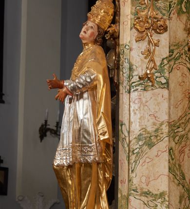 Zdjęcie nr 10: Rzeźba drążona i ścięta, ustawiona na podstawie na planie prostokąta. Święty przedstawiony został w całej postaci, frontalnie, z łukowato wygiętą sylwetką, w kontrapoście, z lewą nogą odciążoną. Obie ręce ma złożone na piersi. Głowę ma uniesioną i zwróconą delikatnie w lewo, twarz podłużną, o łagodnych rysach, wznosi uniesiony, nos wąski, usta grube, zamknięte, na policzkach zaznaczone rumieńce, broda silnie wyodrębniona, zaczerwieniona; głowa okolona krótkimi, kręconymi, brązowymi włosami. Jest ubrany w złotą albę, srebrną komżę dekorowaną u dołu pasem koronki oraz muncet, spod szat wystają czubki złotych butów. Na piersi ma płaskorzeźbione, gorejące serce. Alba i kapa kształtowane sztywno, komża drobnymi fałdami przylega do spodniego ubrania. Partie ciała są polichromowane naturalistyczne. 