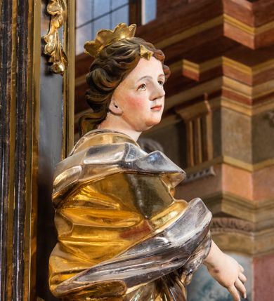 Zdjęcie nr 1: Rzeźba drążona. Ukazana w kontrapoście, ciężar rzeźby opiera się na prawej nodze, lewa zgięta w kolanie i odsunięta lekko w bok. Ręce zgięte w łokciach, umieszczone na wysokości pasa. Głowa obrócona w prawą stronę. Twarz gładka, okrągła, kręcone włosy symetrycznie rozpuszczone po obu stronach twarzy, opadające na plecy. Na głowie umieszczona otwarta korona. Święta odziana jest w długą szatę z głęboko opracowanymi fałdami. Wewnętrzna część szaty jest w kolorze srebrnym, a zewnętrzna jest złocona. Stopy są przyodziane w sandały. Przy lewej nodze świętej umieszczona wieża z oknem oraz krenelażem w zwieńczeniu. 