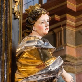 Zdjęcie nr 1: Rzeźba drążona. Ukazana w kontrapoście, ciężar rzeźby opiera się na prawej nodze, lewa zgięta w kolanie i odsunięta lekko w bok. Ręce zgięte w łokciach, umieszczone na wysokości pasa. Głowa obrócona w prawą stronę. Twarz gładka, okrągła, kręcone włosy symetrycznie rozpuszczone po obu stronach twarzy, opadające na plecy. Na głowie umieszczona otwarta korona. Święta odziana jest w długą szatę z głęboko opracowanymi fałdami. Wewnętrzna część szaty jest w kolorze srebrnym, a zewnętrzna jest złocona. Stopy są przyodziane w sandały. Przy lewej nodze świętej umieszczona wieża z oknem oraz krenelażem w zwieńczeniu. 