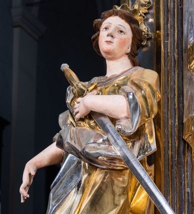 Zdjęcie nr 1: Rzeźba drążona. Święta Katarzyna została ukazana w kontrapoście. Ciężar ciała oparty na prawej nodze, lewa ugięta w kolanie i lekko odsunięta w bok.  Prawa ręka wyprostowana, wyciągnięta w dół pod kątem ok. 45 stopni, lewa ręka zgięta, dłoń dociska miecz do klatki piersiowej.  Twarz okrągła, o rysach młodej kobiety, okolona krótkimi brązowymi włosami spływającymi na plecy. Głowa uniesiona lekko do góry, obrócona nieznacznie w lewą stronę, ozdobiona małą, otwartą koroną. Głownia miecza srebrna, rękojeść złocona. Szata silnie fałdowana, szczególne w okolicy pasa, rękawy podwinięte na wysokość łokci. Stopy odziane w sandały. U stóp świętej umieszczone przerwane koło w kolorze srebrnym.