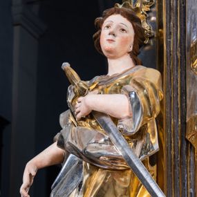 Zdjęcie nr 1: Rzeźba drążona. Święta Katarzyna została ukazana w kontrapoście. Ciężar ciała oparty na prawej nodze, lewa ugięta w kolanie i lekko odsunięta w bok.  Prawa ręka wyprostowana, wyciągnięta w dół pod kątem ok. 45 stopni, lewa ręka zgięta, dłoń dociska miecz do klatki piersiowej.  Twarz okrągła, o rysach młodej kobiety, okolona krótkimi brązowymi włosami spływającymi na plecy. Głowa uniesiona lekko do góry, obrócona nieznacznie w lewą stronę, ozdobiona małą, otwartą koroną. Głownia miecza srebrna, rękojeść złocona. Szata silnie fałdowana, szczególne w okolicy pasa, rękawy podwinięte na wysokość łokci. Stopy odziane w sandały. U stóp świętej umieszczone przerwane koło w kolorze srebrnym.