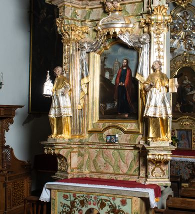 Zdjęcie nr 2: Ołtarz drewniany, prostopadłościenny z kamienną mensą, z kamiennym portatylem. Antepedium drewniane ujęte marmoryzowaną ramą z karbowanym profilem. W polu owalny medalion z przedstawieniem świętego z zakonu Marków, ujętym wicią akantową przeplatana z kwiatową. Nastawa ołtarzowa architektoniczna, jednoosiowa, jednokondygnacyjna ze zwieńczeniem na cokole.  Retabulum flankowane parą zdwojonych półfilarów zakończonych konsolami,  dźwigających pełne belkowanie oraz fragmenty przerwanego, półkolistego przyczółka z siedzącymi puttami. Na tle pilastrów, na konsolach ustawione figury dwóch świętych. Trzony pilastrów dekorowane kampanulami. W polu środkowym wnęka w formie stojącego prostokąta zamkniętego łukiem segmentowym nadwieszonego z uskokiem ujęta profilowaną ramą, z obrazem Wskrzeszenie Łazarza, w osobnej ramie. Nad wnęką baldachim dekorowany lambrekinem, z podwieszoną tkaniną po dwóch stronach, lamowaną frędzlą. Nam baldachimem kartusz z uskrzydlonymi główkami anielskimi i zwieńczony koroną. Zwieńczenie w formie zbliżonej do trapezu o wklęsłych bokach zamknięte falistym gzymsem, dekorowanym kampanulami. W polu gorejące serce przebite mieczem, w otoku z obłoków i uskrzydlonych główek anielskich, w glorii promienistej.  Struktura polichromowana w sposób imitujący marmoryzację w kolorze beżowo-zielonym. Ornamentyka i profile złocone, detal i zwieńczenie srebrzone.