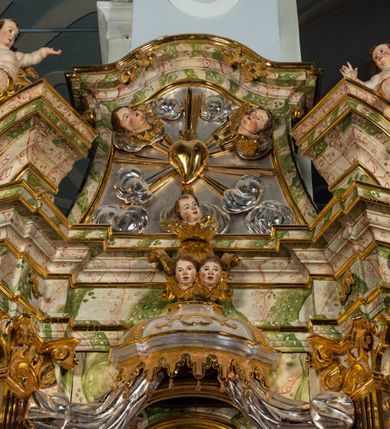 Zdjęcie nr 35: Ołtarz drewniany, prostopadłościenny z kamienną mensą, z kamiennym portatylem. Antepedium drewniane ujęte marmoryzowaną ramą z karbowanym profilem. W polu owalny medalion z przedstawieniem świętego z zakonu Marków, ujętym wicią akantową przeplatana z kwiatową. Nastawa ołtarzowa architektoniczna, jednoosiowa, jednokondygnacyjna ze zwieńczeniem na cokole.  Retabulum flankowane parą zdwojonych półfilarów zakończonych konsolami,  dźwigających pełne belkowanie oraz fragmenty przerwanego, półkolistego przyczółka z siedzącymi puttami. Na tle pilastrów, na konsolach ustawione figury dwóch świętych. Trzony pilastrów dekorowane kampanulami. W polu środkowym wnęka w formie stojącego prostokąta zamkniętego łukiem segmentowym nadwieszonego z uskokiem ujęta profilowaną ramą, z obrazem Wskrzeszenie Łazarza, w osobnej ramie. Nad wnęką baldachim dekorowany lambrekinem, z podwieszoną tkaniną po dwóch stronach, lamowaną frędzlą. Nam baldachimem kartusz z uskrzydlonymi główkami anielskimi i zwieńczony koroną. Zwieńczenie w formie zbliżonej do trapezu o wklęsłych bokach zamknięte falistym gzymsem, dekorowanym kampanulami. W polu gorejące serce przebite mieczem, w otoku z obłoków i uskrzydlonych główek anielskich, w glorii promienistej.  Struktura polichromowana w sposób imitujący marmoryzację w kolorze beżowo-zielonym. Ornamentyka i profile złocone, detal i zwieńczenie srebrzone.