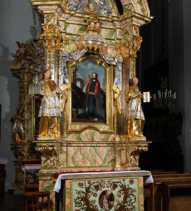 Zdjęcie nr 24: Ołtarz drewniany, prostopadłościenny z kamienną mensą, z kamiennym portatylem. Antepedium drewniane ujęte marmoryzowaną ramą z karbowanym profilem. W polu owalny medalion z przedstawieniem świętego z zakonu Marków, ujętym wicią akantową przeplatana z kwiatową. Nastawa ołtarzowa architektoniczna, jednoosiowa, jednokondygnacyjna ze zwieńczeniem na cokole.  Retabulum flankowane parą zdwojonych półfilarów zakończonych konsolami,  dźwigających pełne belkowanie oraz fragmenty przerwanego, półkolistego przyczółka z siedzącymi puttami. Na tle pilastrów, na konsolach ustawione figury dwóch świętych. Trzony pilastrów dekorowane kampanulami. W polu środkowym wnęka w formie stojącego prostokąta zamkniętego łukiem segmentowym nadwieszonego z uskokiem ujęta profilowaną ramą, z obrazem Wskrzeszenie Łazarza, w osobnej ramie. Nad wnęką baldachim dekorowany lambrekinem, z podwieszoną tkaniną po dwóch stronach, lamowaną frędzlą. Nam baldachimem kartusz z uskrzydlonymi główkami anielskimi i zwieńczony koroną. Zwieńczenie w formie zbliżonej do trapezu o wklęsłych bokach zamknięte falistym gzymsem, dekorowanym kampanulami. W polu gorejące serce przebite mieczem, w otoku z obłoków i uskrzydlonych główek anielskich, w glorii promienistej.  Struktura polichromowana w sposób imitujący marmoryzację w kolorze beżowo-zielonym. Ornamentyka i profile złocone, detal i zwieńczenie srebrzone.