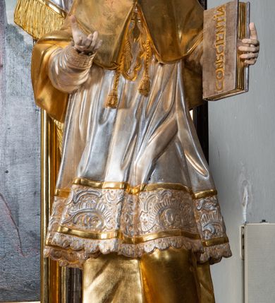 Zdjęcie nr 23: Ołtarz drewniany, prostopadłościenny z kamienną mensą, z kamiennym portatylem. Antepedium drewniane ujęte marmoryzowaną ramą z karbowanym profilem. W polu owalny medalion z przedstawieniem świętego z zakonu Marków, ujętym wicią akantową przeplatana z kwiatową. Nastawa ołtarzowa architektoniczna, jednoosiowa, jednokondygnacyjna ze zwieńczeniem na cokole.  Retabulum flankowane parą zdwojonych półfilarów zakończonych konsolami,  dźwigających pełne belkowanie oraz fragmenty przerwanego, półkolistego przyczółka z siedzącymi puttami. Na tle pilastrów, na konsolach ustawione figury dwóch świętych. Trzony pilastrów dekorowane kampanulami. W polu środkowym wnęka w formie stojącego prostokąta zamkniętego łukiem segmentowym nadwieszonego z uskokiem ujęta profilowaną ramą, z obrazem Wskrzeszenie Łazarza, w osobnej ramie. Nad wnęką baldachim dekorowany lambrekinem, z podwieszoną tkaniną po dwóch stronach, lamowaną frędzlą. Nam baldachimem kartusz z uskrzydlonymi główkami anielskimi i zwieńczony koroną. Zwieńczenie w formie zbliżonej do trapezu o wklęsłych bokach zamknięte falistym gzymsem, dekorowanym kampanulami. W polu gorejące serce przebite mieczem, w otoku z obłoków i uskrzydlonych główek anielskich, w glorii promienistej.  Struktura polichromowana w sposób imitujący marmoryzację w kolorze beżowo-zielonym. Ornamentyka i profile złocone, detal i zwieńczenie srebrzone.