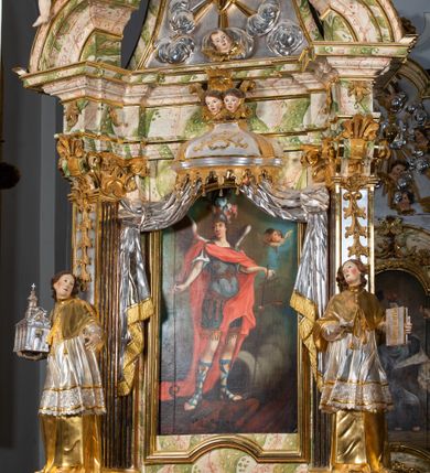 Zdjęcie nr 59: Ołtarz drewniany, prostopadłościenny z kamienną mensą, z kamiennym portatylem. Antepedium drewniane ujęte marmoryzowaną ramą z karbowanym profilem. W polu owalny medalion z przedstawieniem świętego z zakonu Marków, ujętym wicią akantową przeplatana z kwiatową. Nastawa ołtarzowa architektoniczna, jednoosiowa, jednokondygnacyjna ze zwieńczeniem na cokole.  Retabulum flankowane parą zdwojonych półfilarów zakończonych konsolami,  dźwigających pełne belkowanie oraz fragmenty przerwanego, półkolistego przyczółka z siedzącymi puttami. Na tle pilastrów, na konsolach ustawione figury dwóch świętych. Trzony pilastrów dekorowane kampanulami. W polu środkowym wnęka w formie stojącego prostokąta zamkniętego łukiem segmentowym nadwieszonego z uskokiem ujęta profilowaną ramą, z obrazem Wskrzeszenie Łazarza, w osobnej ramie. Nad wnęką baldachim dekorowany lambrekinem, z podwieszoną tkaniną po dwóch stronach, lamowaną frędzlą. Nam baldachimem kartusz z uskrzydlonymi główkami anielskimi i zwieńczony koroną. Zwieńczenie w formie zbliżonej do trapezu o wklęsłych bokach zamknięte falistym gzymsem, dekorowanym kampanulami. W polu gorejące serce przebite mieczem, w otoku z obłoków i uskrzydlonych główek anielskich, w glorii promienistej.  Struktura polichromowana w sposób imitujący marmoryzację w kolorze beżowo-zielonym. Ornamentyka i profile złocone, detal i zwieńczenie srebrzone.