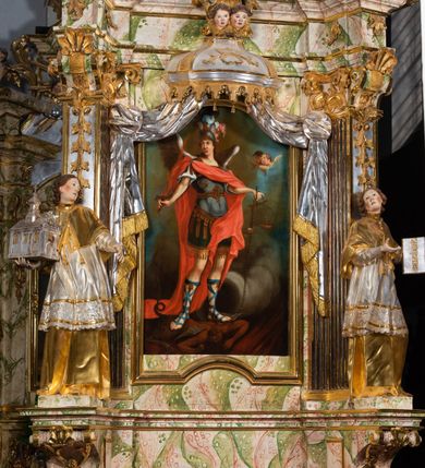 Zdjęcie nr 58: Ołtarz drewniany, prostopadłościenny z kamienną mensą, z kamiennym portatylem. Antepedium drewniane ujęte marmoryzowaną ramą z karbowanym profilem. W polu owalny medalion z przedstawieniem świętego z zakonu Marków, ujętym wicią akantową przeplatana z kwiatową. Nastawa ołtarzowa architektoniczna, jednoosiowa, jednokondygnacyjna ze zwieńczeniem na cokole.  Retabulum flankowane parą zdwojonych półfilarów zakończonych konsolami,  dźwigających pełne belkowanie oraz fragmenty przerwanego, półkolistego przyczółka z siedzącymi puttami. Na tle pilastrów, na konsolach ustawione figury dwóch świętych. Trzony pilastrów dekorowane kampanulami. W polu środkowym wnęka w formie stojącego prostokąta zamkniętego łukiem segmentowym nadwieszonego z uskokiem ujęta profilowaną ramą, z obrazem Wskrzeszenie Łazarza, w osobnej ramie. Nad wnęką baldachim dekorowany lambrekinem, z podwieszoną tkaniną po dwóch stronach, lamowaną frędzlą. Nam baldachimem kartusz z uskrzydlonymi główkami anielskimi i zwieńczony koroną. Zwieńczenie w formie zbliżonej do trapezu o wklęsłych bokach zamknięte falistym gzymsem, dekorowanym kampanulami. W polu gorejące serce przebite mieczem, w otoku z obłoków i uskrzydlonych główek anielskich, w glorii promienistej.  Struktura polichromowana w sposób imitujący marmoryzację w kolorze beżowo-zielonym. Ornamentyka i profile złocone, detal i zwieńczenie srebrzone.