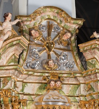 Zdjęcie nr 56: Ołtarz drewniany, prostopadłościenny z kamienną mensą, z kamiennym portatylem. Antepedium drewniane ujęte marmoryzowaną ramą z karbowanym profilem. W polu owalny medalion z przedstawieniem świętego z zakonu Marków, ujętym wicią akantową przeplatana z kwiatową. Nastawa ołtarzowa architektoniczna, jednoosiowa, jednokondygnacyjna ze zwieńczeniem na cokole.  Retabulum flankowane parą zdwojonych półfilarów zakończonych konsolami,  dźwigających pełne belkowanie oraz fragmenty przerwanego, półkolistego przyczółka z siedzącymi puttami. Na tle pilastrów, na konsolach ustawione figury dwóch świętych. Trzony pilastrów dekorowane kampanulami. W polu środkowym wnęka w formie stojącego prostokąta zamkniętego łukiem segmentowym nadwieszonego z uskokiem ujęta profilowaną ramą, z obrazem Wskrzeszenie Łazarza, w osobnej ramie. Nad wnęką baldachim dekorowany lambrekinem, z podwieszoną tkaniną po dwóch stronach, lamowaną frędzlą. Nam baldachimem kartusz z uskrzydlonymi główkami anielskimi i zwieńczony koroną. Zwieńczenie w formie zbliżonej do trapezu o wklęsłych bokach zamknięte falistym gzymsem, dekorowanym kampanulami. W polu gorejące serce przebite mieczem, w otoku z obłoków i uskrzydlonych główek anielskich, w glorii promienistej.  Struktura polichromowana w sposób imitujący marmoryzację w kolorze beżowo-zielonym. Ornamentyka i profile złocone, detal i zwieńczenie srebrzone.