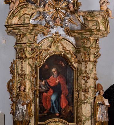 Zdjęcie nr 15: Ołtarz drewniany, prostopadłościenny z kamienną mensą, z kamiennym portatylem. Antependium z prostokątną płyciną ujętą marmoryzowaną ramą ze złoconym karbowanym profilem. W polu medalion w formie stojącego owalu z przedstawieniem zakonnika z dzieciątkiem, flankowany akantową wicią akantową przeplataną z roślinną, na jasnoniebieskim tle. Nastawa ołtarzowa architektoniczna, jednoosiowa, jednokondygnacyjna ze zwieńczeniem na dwustrefowym cokole.  Retabulum flankowane parą opilastrowanych filarów, które dźwigają imposty oraz fragmenty przerwanego półkolistego przyczółka z siedzącymi puttami. Na tle filarów, na konsolach ustawione 2 figury papieża i biskupa. Trzony pilastrów dekorowane zwisami kwiatowymi. W Polu środkowym wnęka w formie stojącego prostokąta zamkniętego łukiem segmentowym nadwieszonego z uskokiem ujęta profilowaną ramą, dekorowaną ornamentem roślinnym, z obrazem świętego Marka. Wnęka od dołu i góry flankowana ornamentem roślinnym i roccailem. Kondygnacja flankowana uszami utworzonymi z roccailu. Zwieńczenie w formie zbliżonej do trapezu o wklęsłych bokach zamknięte fragmentem gzymsu, wyłamanego na kształt łuku segmentowego nadwieszonego, z kartuszem z ornamentem roccailowym. W polu gorejące serce w otoku z obłoków i uskrzydlonych główek anielskich, w glorii promienistej.  Struktura polichromowana w sposób imitujący marmoryzację w kolorze beżowo-zielonym. Ornamentyka i profile złocone, detal i zwieńczenie srebrzone.