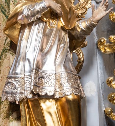 Zdjęcie nr 2: Rzeźba drążona i ścięta, ustawiona na wielobocznej podstawie. Postać ukazana frontalnie z głową skierowaną w prawo, o „esowato” wygiętej sylwetce, w kontrapoście. Prawa ręka złożona na piersi, lewa ugięta i wyciągnięta w bok. Twarz pociągła, owalna, o małych oczach skierowanych do góry, małym zadartym nosie i rozchylonych ustach, z wyodrębnioną brodą, okolona krótkimi, kręconymi, brązowymi włosami. Święty jest ubrany w złotą albę, srebrną komżę z płaskorzeźbioną koronką przy brzegach, a na ramiona ma narzucony złoty mucet zapięty z przodu na guziki, spod szat wystają czubki złotych butów Partie ciała są polichromowane naturalistyczne. 