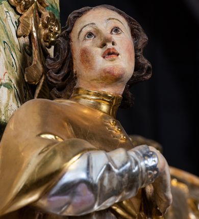 Zdjęcie nr 7: Rzeźba drążona i ścięta, ustawiona na wielobocznej podstawie. Postać ukazana frontalnie z głową skierowaną w prawo, o „esowato” wygiętej sylwetce, w kontrapoście. Prawa ręka złożona na piersi, lewa ugięta i wyciągnięta w bok. Twarz pociągła, owalna, o małych oczach skierowanych do góry, małym zadartym nosie i rozchylonych ustach, z wyodrębnioną brodą, okolona krótkimi, kręconymi, brązowymi włosami. Święty jest ubrany w złotą albę, srebrną komżę z płaskorzeźbioną koronką przy brzegach, a na ramiona ma narzucony złoty mucet zapięty z przodu na guziki, spod szat wystają czubki złotych butów Partie ciała są polichromowane naturalistyczne. 