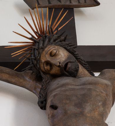 Zdjęcie nr 14: Rzeźba przedstawiająca ukrzyżowanego Chrystusa o ciele ukazanym w delikatnym zwisie ze szczupłymi, wyprężonymi ramionami tworzącymi zarys litery „V”. Głowa Zbawiciela pochylona na prawe ramię z umieszczoną na niej cierniową koroną w formie kołpaka i złoconym nimbem promienistym. Twarz Chrystusa szczupła, oczy zamknięte, nos długi prosty, usta otwarte. Długie, ciemne włosy spływają na prawe ramię długim, skręconym lokiem. Zarost krótki, ciemnobrązowy. Ciało Chrystusa z widoczną raną w prawym boku oddane realistycznie, stopy przebite jednym gwoździem. Wokół bioder udrapowane krótkie, złocone perizonium tworzące płytkie fałdy. U szczytu pionowej belki krzyża titulus w formie banderoli z napisem „INRI”. Krzyż prosty, drewniany w kolorze ciemnobrązowym. Figura w pełni polichromowana, perizonium i nimb złocone.