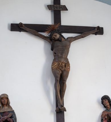Zdjęcie nr 11: Rzeźba przedstawiająca ukrzyżowanego Chrystusa o ciele ukazanym w delikatnym zwisie ze szczupłymi, wyprężonymi ramionami tworzącymi zarys litery „V”. Głowa Zbawiciela pochylona na prawe ramię z umieszczoną na niej cierniową koroną w formie kołpaka i złoconym nimbem promienistym. Twarz Chrystusa szczupła, oczy zamknięte, nos długi prosty, usta otwarte. Długie, ciemne włosy spływają na prawe ramię długim, skręconym lokiem. Zarost krótki, ciemnobrązowy. Ciało Chrystusa z widoczną raną w prawym boku oddane realistycznie, stopy przebite jednym gwoździem. Wokół bioder udrapowane krótkie, złocone perizonium tworzące płytkie fałdy. U szczytu pionowej belki krzyża titulus w formie banderoli z napisem „INRI”. Krzyż prosty, drewniany w kolorze ciemnobrązowym. Figura w pełni polichromowana, perizonium i nimb złocone.