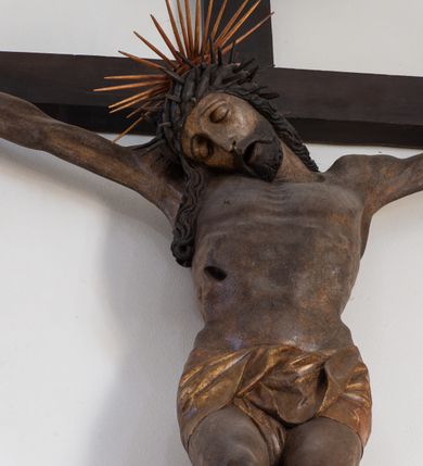 Zdjęcie nr 13: Rzeźba przedstawiająca ukrzyżowanego Chrystusa o ciele ukazanym w delikatnym zwisie ze szczupłymi, wyprężonymi ramionami tworzącymi zarys litery „V”. Głowa Zbawiciela pochylona na prawe ramię z umieszczoną na niej cierniową koroną w formie kołpaka i złoconym nimbem promienistym. Twarz Chrystusa szczupła, oczy zamknięte, nos długi prosty, usta otwarte. Długie, ciemne włosy spływają na prawe ramię długim, skręconym lokiem. Zarost krótki, ciemnobrązowy. Ciało Chrystusa z widoczną raną w prawym boku oddane realistycznie, stopy przebite jednym gwoździem. Wokół bioder udrapowane krótkie, złocone perizonium tworzące płytkie fałdy. U szczytu pionowej belki krzyża titulus w formie banderoli z napisem „INRI”. Krzyż prosty, drewniany w kolorze ciemnobrązowym. Figura w pełni polichromowana, perizonium i nimb złocone.