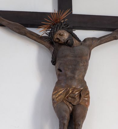Zdjęcie nr 12: Rzeźba przedstawiająca ukrzyżowanego Chrystusa o ciele ukazanym w delikatnym zwisie ze szczupłymi, wyprężonymi ramionami tworzącymi zarys litery „V”. Głowa Zbawiciela pochylona na prawe ramię z umieszczoną na niej cierniową koroną w formie kołpaka i złoconym nimbem promienistym. Twarz Chrystusa szczupła, oczy zamknięte, nos długi prosty, usta otwarte. Długie, ciemne włosy spływają na prawe ramię długim, skręconym lokiem. Zarost krótki, ciemnobrązowy. Ciało Chrystusa z widoczną raną w prawym boku oddane realistycznie, stopy przebite jednym gwoździem. Wokół bioder udrapowane krótkie, złocone perizonium tworzące płytkie fałdy. U szczytu pionowej belki krzyża titulus w formie banderoli z napisem „INRI”. Krzyż prosty, drewniany w kolorze ciemnobrązowym. Figura w pełni polichromowana, perizonium i nimb złocone.