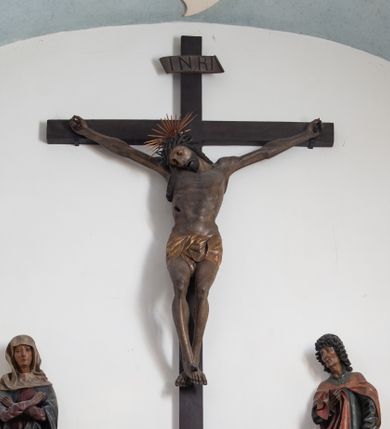 Zdjęcie nr 1: Rzeźba przedstawiająca ukrzyżowanego Chrystusa o ciele ukazanym w delikatnym zwisie ze szczupłymi, wyprężonymi ramionami tworzącymi zarys litery „V”. Głowa Zbawiciela pochylona na prawe ramię z umieszczoną na niej cierniową koroną w formie kołpaka i złoconym nimbem promienistym. Twarz Chrystusa szczupła, oczy zamknięte, nos długi prosty, usta otwarte. Długie, ciemne włosy spływają na prawe ramię długim, skręconym lokiem. Zarost krótki, ciemnobrązowy. Ciało Chrystusa z widoczną raną w prawym boku oddane realistycznie, stopy przebite jednym gwoździem. Wokół bioder udrapowane krótkie, złocone perizonium tworzące płytkie fałdy. U szczytu pionowej belki krzyża titulus w formie banderoli z napisem „INRI”. Krzyż prosty, drewniany w kolorze ciemnobrązowym. Figura w pełni polichromowana, perizonium i nimb złocone.