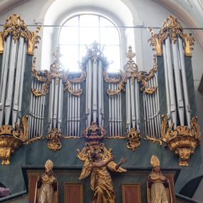 Zdjęcie nr 1: Chór muzyczny konstrukcji drewnianej, wparty na dwóch kamiennych kolumnach o prostych impostowych kapitelach. Balustrada chóru pełna o dekoracji płycinowej (naprzemiennie płyciny owalne, prostokątna i sześcioboczne), zwieńczona profilowanym gzymsem. W dołu ażurowa, wycinana dekoracja. Część środkowa parapetu przełamana i wysunięta do przodu z ustawionymi na niewielkich konsolach polichromowanymi, złoconymi i srebrzonymi figurami: anioła (po środku), św. Wojciecha (?) z lewej i św. Stanisława z prawej. Struktura marmoryzowana w odcieniach zgaszonego niebieskiego, wnętrza płycin w kolorze ceglastej czerwieni. Organy o formie architektonicznej, dziewięcioosiowe z zaakcentowanymi osiami nieparzystymi. Dekorowane złoconym ornamentem akantowym, palmetami, girlandami oraz rocaille'm. Osie trzecia i siódma dodatkowe zwieńczone złoconymi wazonami. Struktura marmoryzowana w odcieniach zgaszonego koloru niebieskiego.