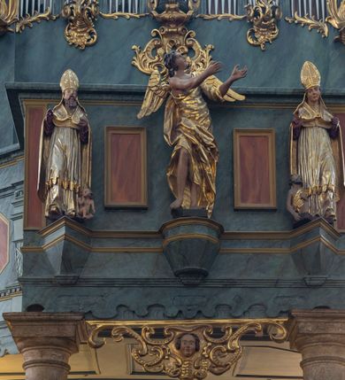 Zdjęcie nr 2: Chór muzyczny konstrukcji drewnianej, wparty na dwóch kamiennych kolumnach o prostych impostowych kapitelach. Balustrada chóru pełna o dekoracji płycinowej (naprzemiennie płyciny owalne, prostokątna i sześcioboczne), zwieńczona profilowanym gzymsem. W dołu ażurowa, wycinana dekoracja. Część środkowa parapetu przełamana i wysunięta do przodu z ustawionymi na niewielkich konsolach polichromowanymi, złoconymi i srebrzonymi figurami: anioła (po środku), św. Wojciecha (?) z lewej i św. Stanisława z prawej. Struktura marmoryzowana w odcieniach zgaszonego niebieskiego, wnętrza płycin w kolorze ceglastej czerwieni. Organy o formie architektonicznej, dziewięcioosiowe z zaakcentowanymi osiami nieparzystymi. Dekorowane złoconym ornamentem akantowym, palmetami, girlandami oraz rocaille'm. Osie trzecia i siódma dodatkowe zwieńczone złoconymi wazonami. Struktura marmoryzowana w odcieniach zgaszonego koloru niebieskiego.