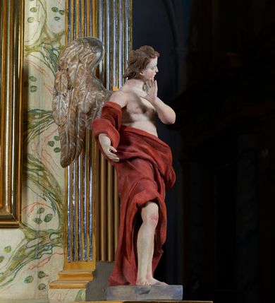 Zdjęcie nr 1: Rzeźba anioła, przyścienna, drewniana, polichromowana, złocona, ścięta i wydrążona od tyłu. Anioł przedstawiony frontalnie, w pozycji stojącej, w kontrapoście. Prawa ręka lekko zgięta wyciągnięta ku przodowi. Lewa dłoń uniesiona ku twarzy. Głowa osadzona na długiej szyi, lekko uniesiona ku górze, przechylona w kierunku prawego ramienia i zwrócona w ¾. Twarz o młodzieńczych rysach, pociągła, dość szczupła, z prostym nosem, drobnymi, lekko rozchylonymi ustami, prostym nosem i wystającym podbródkiem. Głowa okolona jasnobrązowymi, falowanymi wlosami. U ramion anioła skrzydła. Anioł odziany w udrapowane szaty opadające swobodnie wzdłuż ciała i obnażające tors. Polichromia ciała naturalistyczna. Szaty koloru czerwonego. Skrzydła anioła złocone.