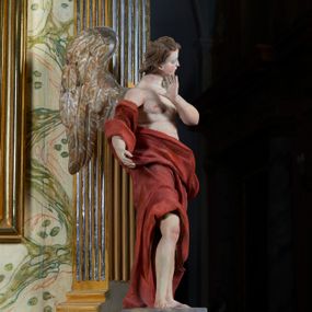 Zdjęcie nr 1: Rzeźba anioła, przyścienna, drewniana, polichromowana, złocona, ścięta i wydrążona od tyłu. Anioł przedstawiony frontalnie, w pozycji stojącej, w kontrapoście. Prawa ręka lekko zgięta wyciągnięta ku przodowi. Lewa dłoń uniesiona ku twarzy. Głowa osadzona na długiej szyi, lekko uniesiona ku górze, przechylona w kierunku prawego ramienia i zwrócona w ¾. Twarz o młodzieńczych rysach, pociągła, dość szczupła, z prostym nosem, drobnymi, lekko rozchylonymi ustami, prostym nosem i wystającym podbródkiem. Głowa okolona jasnobrązowymi, falowanymi wlosami. U ramion anioła skrzydła. Anioł odziany w udrapowane szaty opadające swobodnie wzdłuż ciała i obnażające tors. Polichromia ciała naturalistyczna. Szaty koloru czerwonego. Skrzydła anioła złocone.