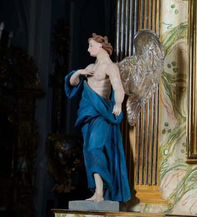 Zdjęcie nr 1: Rzeźba anioła, przyścienna, drewniana, polichromowana, złocona, ścięta i wydrążona od tyłu. Anioł przedstawiony frontalnie, w pozycji stojącej, w kontrapoście. Prawa ręka zgięta w łokciu, uniesiona ku górze, z dłonią przyłożoną do piersi. Lewa ręka lekko odchylona w bok, wyciągnięta wzdłuż ciała. Głowa anioła przechylona w kierunku prawego ramienia, ukazana w ¾. Twarz o młodzieńczych rysach, prostym nosem, rozchylonymi, niewielkimi ustami. Głowa okolona falującymi jasnobrązowymi włosami. U ramion anioła skrzydła. Anioł odziany w udrapowane szaty opadające swobodnie wzdłuż ciała i obnażające tors. Polichromia ciała naturalistyczna. Szaty koloru niebieskiego. Skrzydła złocone.