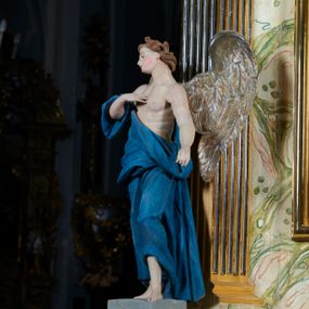 Zdjęcie nr 1: Rzeźba anioła, przyścienna, drewniana, polichromowana, złocona, ścięta i wydrążona od tyłu. Anioł przedstawiony frontalnie, w pozycji stojącej, w kontrapoście. Prawa ręka zgięta w łokciu, uniesiona ku górze, z dłonią przyłożoną do piersi. Lewa ręka lekko odchylona w bok, wyciągnięta wzdłuż ciała. Głowa anioła przechylona w kierunku prawego ramienia, ukazana w ¾. Twarz o młodzieńczych rysach, prostym nosem, rozchylonymi, niewielkimi ustami. Głowa okolona falującymi jasnobrązowymi włosami. U ramion anioła skrzydła. Anioł odziany w udrapowane szaty opadające swobodnie wzdłuż ciała i obnażające tors. Polichromia ciała naturalistyczna. Szaty koloru niebieskiego. Skrzydła złocone.