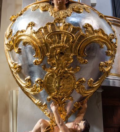 Zdjęcie nr 3: Jest to ambona wisząca, przyścienna, z wejściem od prawej strony. Balustrada na planie półkola, ustawiona na profilowanym cokole, zamknięta silnie wysuniętym gzymsem podtrzymywanym przez pilaster. Ścianka dekorowana płycinami w kształcie leżącego prostokąta po lewej stronie oraz w kształcie stojącego prostokąta po prawej, ozdobione rzeźbionym rocaille’m w polu; schody wąskie, jednobiegowe, połączone ze stojącymi w prezbiterium drewnianymi stallami.  Kosz w kształcie serca, wybrzuszonego w górnej części. U spodu podwieszony anioł z rękami wyciągniętymi do góry i ugiętymi w kolanach nogami, w pasie przewiązany złotą tkaniną, silnie drapowaną. W górnej części kosza, pośrodku, uskrzydlona główka aniołka, skierowana na wprost. Twarz owalna o pulchnych policzkach, z wyraźnie zaznaczonym podbródkiem, usta delikatnie otwarte, nos spiczasty, oczy skierowane ku górze; włosy średniej długości, ciemnobrązowe z loczkiem na czubku głowy. Powierzchnia kosza pokryta płaskorzeźbioną dekoracją rocaille’ową. Zaplecek w kształcie stojącego prostokąta, flankowany dwoma pilastrami, podtrzymującymi przerywany, odwrócony, wolutowy przyczółek. W polu wpisany duży, łaciński krzyż o rozszerzających się ramionach, wysunięty poza boki zaplecka.  Pilastry od wewnątrz zdobione są pasem gęstej  dekoracji rocaille’owej. Nad amboną zawieszona jest gołębica Ducha Świętego na tle promienistej glorii, utworzonej z wiązek promieni o nierównej długości. Na promieniach umieszczone są cztery uskrzydlone główki aniołków. Struktura srebrzona, dekoracja ornamentalna złocona.