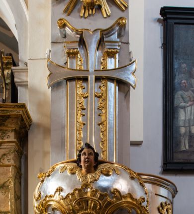 Zdjęcie nr 2: Jest to ambona wisząca, przyścienna, z wejściem od prawej strony. Balustrada na planie półkola, ustawiona na profilowanym cokole, zamknięta silnie wysuniętym gzymsem podtrzymywanym przez pilaster. Ścianka dekorowana płycinami w kształcie leżącego prostokąta po lewej stronie oraz w kształcie stojącego prostokąta po prawej, ozdobione rzeźbionym rocaille’m w polu; schody wąskie, jednobiegowe, połączone ze stojącymi w prezbiterium drewnianymi stallami.  Kosz w kształcie serca, wybrzuszonego w górnej części. U spodu podwieszony anioł z rękami wyciągniętymi do góry i ugiętymi w kolanach nogami, w pasie przewiązany złotą tkaniną, silnie drapowaną. W górnej części kosza, pośrodku, uskrzydlona główka aniołka, skierowana na wprost. Twarz owalna o pulchnych policzkach, z wyraźnie zaznaczonym podbródkiem, usta delikatnie otwarte, nos spiczasty, oczy skierowane ku górze; włosy średniej długości, ciemnobrązowe z loczkiem na czubku głowy. Powierzchnia kosza pokryta płaskorzeźbioną dekoracją rocaille’ową. Zaplecek w kształcie stojącego prostokąta, flankowany dwoma pilastrami, podtrzymującymi przerywany, odwrócony, wolutowy przyczółek. W polu wpisany duży, łaciński krzyż o rozszerzających się ramionach, wysunięty poza boki zaplecka.  Pilastry od wewnątrz zdobione są pasem gęstej  dekoracji rocaille’owej. Nad amboną zawieszona jest gołębica Ducha Świętego na tle promienistej glorii, utworzonej z wiązek promieni o nierównej długości. Na promieniach umieszczone są cztery uskrzydlone główki aniołków. Struktura srebrzona, dekoracja ornamentalna złocona.