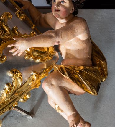 Zdjęcie nr 4: Jest to ambona wisząca, przyścienna, z wejściem od prawej strony. Balustrada na planie półkola, ustawiona na profilowanym cokole, zamknięta silnie wysuniętym gzymsem podtrzymywanym przez pilaster. Ścianka dekorowana płycinami w kształcie leżącego prostokąta po lewej stronie oraz w kształcie stojącego prostokąta po prawej, ozdobione rzeźbionym rocaille’m w polu; schody wąskie, jednobiegowe, połączone ze stojącymi w prezbiterium drewnianymi stallami.  Kosz w kształcie serca, wybrzuszonego w górnej części. U spodu podwieszony anioł z rękami wyciągniętymi do góry i ugiętymi w kolanach nogami, w pasie przewiązany złotą tkaniną, silnie drapowaną. W górnej części kosza, pośrodku, uskrzydlona główka aniołka, skierowana na wprost. Twarz owalna o pulchnych policzkach, z wyraźnie zaznaczonym podbródkiem, usta delikatnie otwarte, nos spiczasty, oczy skierowane ku górze; włosy średniej długości, ciemnobrązowe z loczkiem na czubku głowy. Powierzchnia kosza pokryta płaskorzeźbioną dekoracją rocaille’ową. Zaplecek w kształcie stojącego prostokąta, flankowany dwoma pilastrami, podtrzymującymi przerywany, odwrócony, wolutowy przyczółek. W polu wpisany duży, łaciński krzyż o rozszerzających się ramionach, wysunięty poza boki zaplecka.  Pilastry od wewnątrz zdobione są pasem gęstej  dekoracji rocaille’owej. Nad amboną zawieszona jest gołębica Ducha Świętego na tle promienistej glorii, utworzonej z wiązek promieni o nierównej długości. Na promieniach umieszczone są cztery uskrzydlone główki aniołków. Struktura srebrzona, dekoracja ornamentalna złocona.