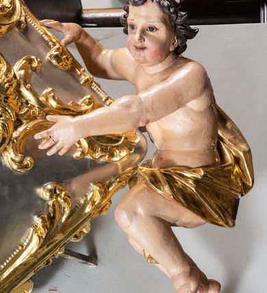 Zdjęcie nr 8: Jest to ambona wisząca, przyścienna, z wejściem od prawej strony. Balustrada na planie półkola, ustawiona na profilowanym cokole, zamknięta silnie wysuniętym gzymsem podtrzymywanym przez pilaster. Ścianka dekorowana płycinami w kształcie leżącego prostokąta po lewej stronie oraz w kształcie stojącego prostokąta po prawej, ozdobione rzeźbionym rocaille’m w polu; schody wąskie, jednobiegowe, połączone ze stojącymi w prezbiterium drewnianymi stallami.  Kosz w kształcie serca, wybrzuszonego w górnej części. U spodu podwieszony anioł z rękami wyciągniętymi do góry i ugiętymi w kolanach nogami, w pasie przewiązany złotą tkaniną, silnie drapowaną. W górnej części kosza, pośrodku, uskrzydlona główka aniołka, skierowana na wprost. Twarz owalna o pulchnych policzkach, z wyraźnie zaznaczonym podbródkiem, usta delikatnie otwarte, nos spiczasty, oczy skierowane ku górze; włosy średniej długości, ciemnobrązowe z loczkiem na czubku głowy. Powierzchnia kosza pokryta płaskorzeźbioną dekoracją rocaille’ową. Zaplecek w kształcie stojącego prostokąta, flankowany dwoma pilastrami, podtrzymującymi przerywany, odwrócony, wolutowy przyczółek. W polu wpisany duży, łaciński krzyż o rozszerzających się ramionach, wysunięty poza boki zaplecka.  Pilastry od wewnątrz zdobione są pasem gęstej  dekoracji rocaille’owej. Nad amboną zawieszona jest gołębica Ducha Świętego na tle promienistej glorii, utworzonej z wiązek promieni o nierównej długości. Na promieniach umieszczone są cztery uskrzydlone główki aniołków. Struktura srebrzona, dekoracja ornamentalna złocona.