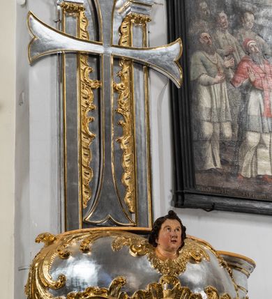 Zdjęcie nr 6: Jest to ambona wisząca, przyścienna, z wejściem od prawej strony. Balustrada na planie półkola, ustawiona na profilowanym cokole, zamknięta silnie wysuniętym gzymsem podtrzymywanym przez pilaster. Ścianka dekorowana płycinami w kształcie leżącego prostokąta po lewej stronie oraz w kształcie stojącego prostokąta po prawej, ozdobione rzeźbionym rocaille’m w polu; schody wąskie, jednobiegowe, połączone ze stojącymi w prezbiterium drewnianymi stallami.  Kosz w kształcie serca, wybrzuszonego w górnej części. U spodu podwieszony anioł z rękami wyciągniętymi do góry i ugiętymi w kolanach nogami, w pasie przewiązany złotą tkaniną, silnie drapowaną. W górnej części kosza, pośrodku, uskrzydlona główka aniołka, skierowana na wprost. Twarz owalna o pulchnych policzkach, z wyraźnie zaznaczonym podbródkiem, usta delikatnie otwarte, nos spiczasty, oczy skierowane ku górze; włosy średniej długości, ciemnobrązowe z loczkiem na czubku głowy. Powierzchnia kosza pokryta płaskorzeźbioną dekoracją rocaille’ową. Zaplecek w kształcie stojącego prostokąta, flankowany dwoma pilastrami, podtrzymującymi przerywany, odwrócony, wolutowy przyczółek. W polu wpisany duży, łaciński krzyż o rozszerzających się ramionach, wysunięty poza boki zaplecka.  Pilastry od wewnątrz zdobione są pasem gęstej  dekoracji rocaille’owej. Nad amboną zawieszona jest gołębica Ducha Świętego na tle promienistej glorii, utworzonej z wiązek promieni o nierównej długości. Na promieniach umieszczone są cztery uskrzydlone główki aniołków. Struktura srebrzona, dekoracja ornamentalna złocona.