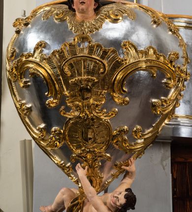 Zdjęcie nr 7: Jest to ambona wisząca, przyścienna, z wejściem od prawej strony. Balustrada na planie półkola, ustawiona na profilowanym cokole, zamknięta silnie wysuniętym gzymsem podtrzymywanym przez pilaster. Ścianka dekorowana płycinami w kształcie leżącego prostokąta po lewej stronie oraz w kształcie stojącego prostokąta po prawej, ozdobione rzeźbionym rocaille’m w polu; schody wąskie, jednobiegowe, połączone ze stojącymi w prezbiterium drewnianymi stallami.  Kosz w kształcie serca, wybrzuszonego w górnej części. U spodu podwieszony anioł z rękami wyciągniętymi do góry i ugiętymi w kolanach nogami, w pasie przewiązany złotą tkaniną, silnie drapowaną. W górnej części kosza, pośrodku, uskrzydlona główka aniołka, skierowana na wprost. Twarz owalna o pulchnych policzkach, z wyraźnie zaznaczonym podbródkiem, usta delikatnie otwarte, nos spiczasty, oczy skierowane ku górze; włosy średniej długości, ciemnobrązowe z loczkiem na czubku głowy. Powierzchnia kosza pokryta płaskorzeźbioną dekoracją rocaille’ową. Zaplecek w kształcie stojącego prostokąta, flankowany dwoma pilastrami, podtrzymującymi przerywany, odwrócony, wolutowy przyczółek. W polu wpisany duży, łaciński krzyż o rozszerzających się ramionach, wysunięty poza boki zaplecka.  Pilastry od wewnątrz zdobione są pasem gęstej  dekoracji rocaille’owej. Nad amboną zawieszona jest gołębica Ducha Świętego na tle promienistej glorii, utworzonej z wiązek promieni o nierównej długości. Na promieniach umieszczone są cztery uskrzydlone główki aniołków. Struktura srebrzona, dekoracja ornamentalna złocona.