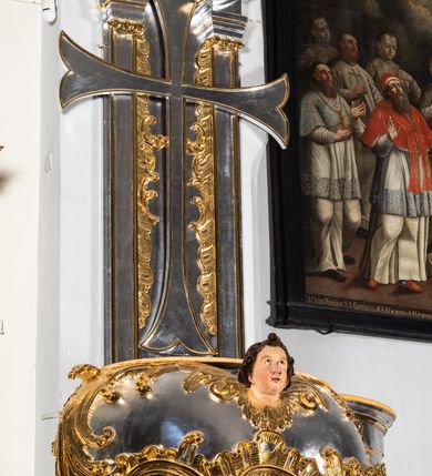 Zdjęcie nr 5: Jest to ambona wisząca, przyścienna, z wejściem od prawej strony. Balustrada na planie półkola, ustawiona na profilowanym cokole, zamknięta silnie wysuniętym gzymsem podtrzymywanym przez pilaster. Ścianka dekorowana płycinami w kształcie leżącego prostokąta po lewej stronie oraz w kształcie stojącego prostokąta po prawej, ozdobione rzeźbionym rocaille’m w polu; schody wąskie, jednobiegowe, połączone ze stojącymi w prezbiterium drewnianymi stallami.  Kosz w kształcie serca, wybrzuszonego w górnej części. U spodu podwieszony anioł z rękami wyciągniętymi do góry i ugiętymi w kolanach nogami, w pasie przewiązany złotą tkaniną, silnie drapowaną. W górnej części kosza, pośrodku, uskrzydlona główka aniołka, skierowana na wprost. Twarz owalna o pulchnych policzkach, z wyraźnie zaznaczonym podbródkiem, usta delikatnie otwarte, nos spiczasty, oczy skierowane ku górze; włosy średniej długości, ciemnobrązowe z loczkiem na czubku głowy. Powierzchnia kosza pokryta płaskorzeźbioną dekoracją rocaille’ową. Zaplecek w kształcie stojącego prostokąta, flankowany dwoma pilastrami, podtrzymującymi przerywany, odwrócony, wolutowy przyczółek. W polu wpisany duży, łaciński krzyż o rozszerzających się ramionach, wysunięty poza boki zaplecka.  Pilastry od wewnątrz zdobione są pasem gęstej  dekoracji rocaille’owej. Nad amboną zawieszona jest gołębica Ducha Świętego na tle promienistej glorii, utworzonej z wiązek promieni o nierównej długości. Na promieniach umieszczone są cztery uskrzydlone główki aniołków. Struktura srebrzona, dekoracja ornamentalna złocona.