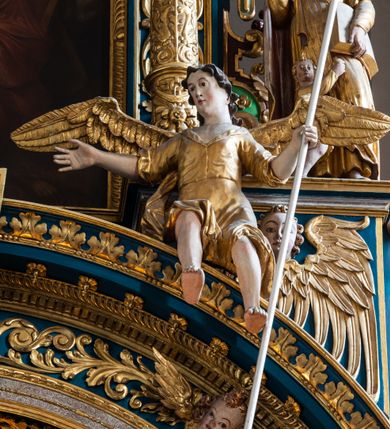 Zdjęcie nr 37: Ołtarz architektoniczny, trójosiowy, dwukondygnacyjny ze zwieńczeniem. Mensa prostopadłościenna, kamienna z drewnianym antepedium w formie leżącego prostokąta z postacią św. Augustyna ujętego wicią suchego akantu z przeplecioną, karbowaną wstążką i bukietami kwiatów. Przedstawienie otoczone profilowaną, ażurową ramą wypełnioną podobną wicią z akantu i kwiatów. Na mensie prostopadłościenne tabernakulum w formie kapliczki. Ściana frontowa z drzwiczkami w kształcie stojącego prostokąta dekorowanego krzyżem łacińskim ujętym wicią roślinną, zwieńczona trójkątnym przyczółkiem. Ścianka ujęta lizenami wypełnionymi wicią akantową podtrzymującymi pełne belkowanie obiegające bryłę, z mocno wysuniętym gzymsem wyłamanym w narożach. Całość zwieńczona czterospadowym dachem dekorowanym łuską. Na dachu baranek Boży leżący na księdze z siedmioma pieczęciami, przytrzymujący chorągiew, ukazany na tle chrystogramu otoczonego glorią promienistą. 
Nastawa na dwudzielnym cokole z półkolistymi bramkami ze zwieńczeniem utworzonym z ornamentu okuciowego ujmującego owalne pola. Osie pierwszej kondygnacji wydzielone kolumnami stojącymi na konsolach dekorowanych z frontu uskrzydlonymi główkami anielskimi powyżej liści akantu, na krawędziach cekinami, a po bokach wicią. Kolumny zostały przedzielone opaską w jednej trzeciej wysokości, dekorowane na całej wysokości wicią roślinną, główkami anielskimi w dolnej partii i imitacją kameryzacji na opaskach oraz w dwóch trzecich wysokości. Kolumny są ustawione na tle pilastrów dekorowanych wicią roślinną i zawieszonymi na niej chustkami. W polu centralnym kondygnacji jest nisza w kształcie stojącego prostokąta zamkniętego łukiem pełnym z postacią ukrzyżowanego Chrystusa na tle widoku Jerozolimy. W osiach bocznych mniejsze, ujęte pilastrami nisze o podobnym kształcie, których konchy wypełnione są muszlą, zwieńczone trójkątnym przyczółkiem przerwanym na cokół. W nich, na dekorowanych wicią i rozetami konsolach ustawiono figury: po lewej stronie św. Piotra, a po prawej św. Pawła. Kondygnacje ujmują ażurowe uszaki utworzone z ornamentu okuciowego, ulistnionych wolut i przewieszonej chustki. Kolumny podtrzymują pełne belkowanie wyniesione w łuk nad polem centralnym. Podłucze dekorowane rozetami, fryz głowami anielskimi i wicią, gzyms m.in. kostkowaniem i liśćmi akantu. Całość spięta kluczem z kartuszem z herbem kanoników regularnych od pokuty (marków). Na wyniesieniu belkowania posadowiono postaci aniołów trzymających narzędzia męki: anioł po lewej włócznię, a anioł po prawej gąbkę. Górna kondygnacja oparta została na schemacie aediculi – prostokątne pole ujęto kolumnami podtrzymującymi pełne belkowanie i trójkątny przyczółek przerwany na środku na cokół, na którym ustawiono rzeźbę  pelikana karmiącego młode własną krwią. Na krawędziach przyczółka dwie leżące postaci. Kondygnacja flankowana postaciami czterech ewangelistów ustawionymi na cokołach. Po lewej stronie św. Marek i św. Jan, po prawej św. Mateusz i św. Łukasz. Struktura polichromowana na kolor ciemnoniebieski; kolumny, profile, detale i ornamenty złocone i srebrzone.
