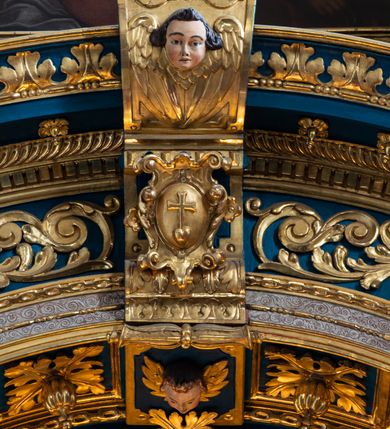 Zdjęcie nr 38: Ołtarz architektoniczny, trójosiowy, dwukondygnacyjny ze zwieńczeniem. Mensa prostopadłościenna, kamienna z drewnianym antepedium w formie leżącego prostokąta z postacią św. Augustyna ujętego wicią suchego akantu z przeplecioną, karbowaną wstążką i bukietami kwiatów. Przedstawienie otoczone profilowaną, ażurową ramą wypełnioną podobną wicią z akantu i kwiatów. Na mensie prostopadłościenne tabernakulum w formie kapliczki. Ściana frontowa z drzwiczkami w kształcie stojącego prostokąta dekorowanego krzyżem łacińskim ujętym wicią roślinną, zwieńczona trójkątnym przyczółkiem. Ścianka ujęta lizenami wypełnionymi wicią akantową podtrzymującymi pełne belkowanie obiegające bryłę, z mocno wysuniętym gzymsem wyłamanym w narożach. Całość zwieńczona czterospadowym dachem dekorowanym łuską. Na dachu baranek Boży leżący na księdze z siedmioma pieczęciami, przytrzymujący chorągiew, ukazany na tle chrystogramu otoczonego glorią promienistą. 
Nastawa na dwudzielnym cokole z półkolistymi bramkami ze zwieńczeniem utworzonym z ornamentu okuciowego ujmującego owalne pola. Osie pierwszej kondygnacji wydzielone kolumnami stojącymi na konsolach dekorowanych z frontu uskrzydlonymi główkami anielskimi powyżej liści akantu, na krawędziach cekinami, a po bokach wicią. Kolumny zostały przedzielone opaską w jednej trzeciej wysokości, dekorowane na całej wysokości wicią roślinną, główkami anielskimi w dolnej partii i imitacją kameryzacji na opaskach oraz w dwóch trzecich wysokości. Kolumny są ustawione na tle pilastrów dekorowanych wicią roślinną i zawieszonymi na niej chustkami. W polu centralnym kondygnacji jest nisza w kształcie stojącego prostokąta zamkniętego łukiem pełnym z postacią ukrzyżowanego Chrystusa na tle widoku Jerozolimy. W osiach bocznych mniejsze, ujęte pilastrami nisze o podobnym kształcie, których konchy wypełnione są muszlą, zwieńczone trójkątnym przyczółkiem przerwanym na cokół. W nich, na dekorowanych wicią i rozetami konsolach ustawiono figury: po lewej stronie św. Piotra, a po prawej św. Pawła. Kondygnacje ujmują ażurowe uszaki utworzone z ornamentu okuciowego, ulistnionych wolut i przewieszonej chustki. Kolumny podtrzymują pełne belkowanie wyniesione w łuk nad polem centralnym. Podłucze dekorowane rozetami, fryz głowami anielskimi i wicią, gzyms m.in. kostkowaniem i liśćmi akantu. Całość spięta kluczem z kartuszem z herbem kanoników regularnych od pokuty (marków). Na wyniesieniu belkowania posadowiono postaci aniołów trzymających narzędzia męki: anioł po lewej włócznię, a anioł po prawej gąbkę. Górna kondygnacja oparta została na schemacie aediculi – prostokątne pole ujęto kolumnami podtrzymującymi pełne belkowanie i trójkątny przyczółek przerwany na środku na cokół, na którym ustawiono rzeźbę  pelikana karmiącego młode własną krwią. Na krawędziach przyczółka dwie leżące postaci. Kondygnacja flankowana postaciami czterech ewangelistów ustawionymi na cokołach. Po lewej stronie św. Marek i św. Jan, po prawej św. Mateusz i św. Łukasz. Struktura polichromowana na kolor ciemnoniebieski; kolumny, profile, detale i ornamenty złocone i srebrzone.
