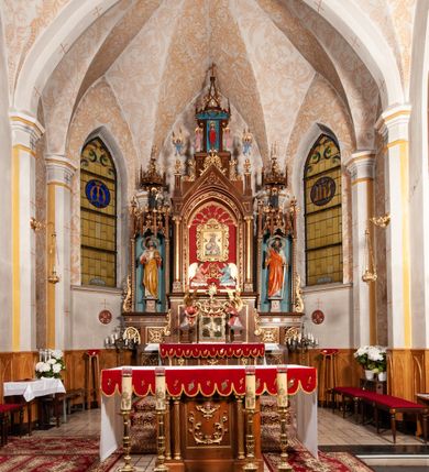 Zdjęcie nr 31: Ołtarz jednokondygnacyjny na wysokim, schodkowym cokole, trójosiowy, z wysuniętą mensą ołtarzową. Płyciny cokołu zdobione dekoracją w formie ostrołukowych maswerków i rozet w kwadratowej ramie. Antepedium flankowane parą kolumn ustawionych na wysokich cokołach i kanelowanych, skręconych trzonach z kompozytowymi kapitelami. W polu dwie prostokątne płyciny i kwadratowa pośrodku ze stylizowaną wicią roślinną i małą uskrzydloną główką aniołka. Tabernakulum proste, w kształcie prostopadłościennej skrzynki, o złotych drzwiczkach z wytłaczaną formą równoramiennego krzyża i wypukłą srebrną rybą oraz koszem z chlebem. Po obu stronach, na niewysokich postumentach dwa klęczące anioły z wysokimi skrzydłami, w biało różowych szatach z fioletowym podbiciem (skrzydła stylizowane na husarskie). Anioł po lewej z kadzielnicą, a anioł po prawej z kielichem.
Nastawa ołtarzowa o trzech oddzielnych niszach, nakrytych gotycyzującymi baldachimami z pinaklami, ozdobionymi żabkami. Oś środkowa w formie niszy zamkniętej ostrołukowo z wimpergą wspartą na smukłych, złoconych kolumienkach o kapitelach kompozytowych. Wimperga ze złoconymi, profilowanymi płycinami, zwieńczona rzeźbionymi liśćmi akantu i wolutami. Na przedłużeniu kolumn postumenty z figurami aniołów w biało niebieskich szatach, ze złotymi, wydłużonymi skrzydłami. Na osi na postumencie, pod gotycyzującym baldachimem figura świętego biskupa w czerwonej szacie z krzyżem w dłoni pomiędzy dwoma aniołami w białych i różowych szatach. W niszy, na czerwonym tle obraz Matki Boskiej Nieustającej Pomocy w prostej ramie z dekoracją w formie wici roślinnej. Poniżej dwa płaskorzeźbione anioły trzymające ażurowy monogram maryjny. Anioły o pozłacanych skrzydłach, na chmurach, w różowej i niebieskiej szacie. Osie boczne w formie aediculi z gotycyzującym baldachimem. W niszach figury, po lewej św. Piotra, a po prawej św. Pawła. Aedicule ujęte są smukłymi, skręconymi kolumnami z kapitelami wolutowymi oraz roślinnym uszakiem z perełkowaniem. Nad nimi imposty, na których rzeźby osłaniane baldachimami, po lewej świętego w habicie jezuity, a po prawej św. Jana Kantego. Drewno bejcowane na ciemny brąz, z nakładanymi elementami dekoracyjnymi złoconymi. 