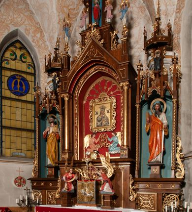 Zdjęcie nr 32: Ołtarz jednokondygnacyjny na wysokim, schodkowym cokole, trójosiowy, z wysuniętą mensą ołtarzową. Płyciny cokołu zdobione dekoracją w formie ostrołukowych maswerków i rozet w kwadratowej ramie. Antepedium flankowane parą kolumn ustawionych na wysokich cokołach i kanelowanych, skręconych trzonach z kompozytowymi kapitelami. W polu dwie prostokątne płyciny i kwadratowa pośrodku ze stylizowaną wicią roślinną i małą uskrzydloną główką aniołka. Tabernakulum proste, w kształcie prostopadłościennej skrzynki, o złotych drzwiczkach z wytłaczaną formą równoramiennego krzyża i wypukłą srebrną rybą oraz koszem z chlebem. Po obu stronach, na niewysokich postumentach dwa klęczące anioły z wysokimi skrzydłami, w biało różowych szatach z fioletowym podbiciem (skrzydła stylizowane na husarskie). Anioł po lewej z kadzielnicą, a anioł po prawej z kielichem.
Nastawa ołtarzowa o trzech oddzielnych niszach, nakrytych gotycyzującymi baldachimami z pinaklami, ozdobionymi żabkami. Oś środkowa w formie niszy zamkniętej ostrołukowo z wimpergą wspartą na smukłych, złoconych kolumienkach o kapitelach kompozytowych. Wimperga ze złoconymi, profilowanymi płycinami, zwieńczona rzeźbionymi liśćmi akantu i wolutami. Na przedłużeniu kolumn postumenty z figurami aniołów w biało niebieskich szatach, ze złotymi, wydłużonymi skrzydłami. Na osi na postumencie, pod gotycyzującym baldachimem figura świętego biskupa w czerwonej szacie z krzyżem w dłoni pomiędzy dwoma aniołami w białych i różowych szatach. W niszy, na czerwonym tle obraz Matki Boskiej Nieustającej Pomocy w prostej ramie z dekoracją w formie wici roślinnej. Poniżej dwa płaskorzeźbione anioły trzymające ażurowy monogram maryjny. Anioły o pozłacanych skrzydłach, na chmurach, w różowej i niebieskiej szacie. Osie boczne w formie aediculi z gotycyzującym baldachimem. W niszach figury, po lewej św. Piotra, a po prawej św. Pawła. Aedicule ujęte są smukłymi, skręconymi kolumnami z kapitelami wolutowymi oraz roślinnym uszakiem z perełkowaniem. Nad nimi imposty, na których rzeźby osłaniane baldachimami, po lewej świętego w habicie jezuity, a po prawej św. Jana Kantego. Drewno bejcowane na ciemny brąz, z nakładanymi elementami dekoracyjnymi złoconymi. 