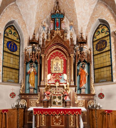Zdjęcie nr 1: Ołtarz jednokondygnacyjny na wysokim, schodkowym cokole, trójosiowy, z wysuniętą mensą ołtarzową. Płyciny cokołu zdobione dekoracją w formie ostrołukowych maswerków i rozet w kwadratowej ramie. Antepedium flankowane parą kolumn ustawionych na wysokich cokołach i kanelowanych, skręconych trzonach z kompozytowymi kapitelami. W polu dwie prostokątne płyciny i kwadratowa pośrodku ze stylizowaną wicią roślinną i małą uskrzydloną główką aniołka. Tabernakulum proste, w kształcie prostopadłościennej skrzynki, o złotych drzwiczkach z wytłaczaną formą równoramiennego krzyża i wypukłą srebrną rybą oraz koszem z chlebem. Po obu stronach, na niewysokich postumentach dwa klęczące anioły z wysokimi skrzydłami, w biało różowych szatach z fioletowym podbiciem (skrzydła stylizowane na husarskie). Anioł po lewej z kadzielnicą, a anioł po prawej z kielichem.
Nastawa ołtarzowa o trzech oddzielnych niszach, nakrytych gotycyzującymi baldachimami z pinaklami, ozdobionymi żabkami. Oś środkowa w formie niszy zamkniętej ostrołukowo z wimpergą wspartą na smukłych, złoconych kolumienkach o kapitelach kompozytowych. Wimperga ze złoconymi, profilowanymi płycinami, zwieńczona rzeźbionymi liśćmi akantu i wolutami. Na przedłużeniu kolumn postumenty z figurami aniołów w biało niebieskich szatach, ze złotymi, wydłużonymi skrzydłami. Na osi na postumencie, pod gotycyzującym baldachimem figura świętego biskupa w czerwonej szacie z krzyżem w dłoni pomiędzy dwoma aniołami w białych i różowych szatach. W niszy, na czerwonym tle obraz Matki Boskiej Nieustającej Pomocy w prostej ramie z dekoracją w formie wici roślinnej. Poniżej dwa płaskorzeźbione anioły trzymające ażurowy monogram maryjny. Anioły o pozłacanych skrzydłach, na chmurach, w różowej i niebieskiej szacie. Osie boczne w formie aediculi z gotycyzującym baldachimem. W niszach figury, po lewej św. Piotra, a po prawej św. Pawła. Aedicule ujęte są smukłymi, skręconymi kolumnami z kapitelami wolutowymi oraz roślinnym uszakiem z perełkowaniem. Nad nimi imposty, na których rzeźby osłaniane baldachimami, po lewej świętego w habicie jezuity, a po prawej św. Jana Kantego. Drewno bejcowane na ciemny brąz, z nakładanymi elementami dekoracyjnymi złoconymi. 
