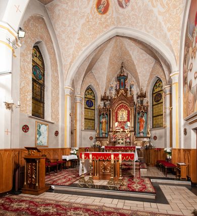 Zdjęcie nr 33: Ołtarz jednokondygnacyjny na wysokim, schodkowym cokole, trójosiowy, z wysuniętą mensą ołtarzową. Płyciny cokołu zdobione dekoracją w formie ostrołukowych maswerków i rozet w kwadratowej ramie. Antepedium flankowane parą kolumn ustawionych na wysokich cokołach i kanelowanych, skręconych trzonach z kompozytowymi kapitelami. W polu dwie prostokątne płyciny i kwadratowa pośrodku ze stylizowaną wicią roślinną i małą uskrzydloną główką aniołka. Tabernakulum proste, w kształcie prostopadłościennej skrzynki, o złotych drzwiczkach z wytłaczaną formą równoramiennego krzyża i wypukłą srebrną rybą oraz koszem z chlebem. Po obu stronach, na niewysokich postumentach dwa klęczące anioły z wysokimi skrzydłami, w biało różowych szatach z fioletowym podbiciem (skrzydła stylizowane na husarskie). Anioł po lewej z kadzielnicą, a anioł po prawej z kielichem.
Nastawa ołtarzowa o trzech oddzielnych niszach, nakrytych gotycyzującymi baldachimami z pinaklami, ozdobionymi żabkami. Oś środkowa w formie niszy zamkniętej ostrołukowo z wimpergą wspartą na smukłych, złoconych kolumienkach o kapitelach kompozytowych. Wimperga ze złoconymi, profilowanymi płycinami, zwieńczona rzeźbionymi liśćmi akantu i wolutami. Na przedłużeniu kolumn postumenty z figurami aniołów w biało niebieskich szatach, ze złotymi, wydłużonymi skrzydłami. Na osi na postumencie, pod gotycyzującym baldachimem figura świętego biskupa w czerwonej szacie z krzyżem w dłoni pomiędzy dwoma aniołami w białych i różowych szatach. W niszy, na czerwonym tle obraz Matki Boskiej Nieustającej Pomocy w prostej ramie z dekoracją w formie wici roślinnej. Poniżej dwa płaskorzeźbione anioły trzymające ażurowy monogram maryjny. Anioły o pozłacanych skrzydłach, na chmurach, w różowej i niebieskiej szacie. Osie boczne w formie aediculi z gotycyzującym baldachimem. W niszach figury, po lewej św. Piotra, a po prawej św. Pawła. Aedicule ujęte są smukłymi, skręconymi kolumnami z kapitelami wolutowymi oraz roślinnym uszakiem z perełkowaniem. Nad nimi imposty, na których rzeźby osłaniane baldachimami, po lewej świętego w habicie jezuity, a po prawej św. Jana Kantego. Drewno bejcowane na ciemny brąz, z nakładanymi elementami dekoracyjnymi złoconymi. 