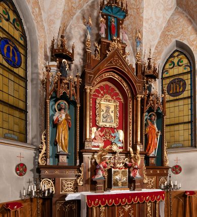 Zdjęcie nr 2: Ołtarz jednokondygnacyjny na wysokim, schodkowym cokole, trójosiowy, z wysuniętą mensą ołtarzową. Płyciny cokołu zdobione dekoracją w formie ostrołukowych maswerków i rozet w kwadratowej ramie. Antepedium flankowane parą kolumn ustawionych na wysokich cokołach i kanelowanych, skręconych trzonach z kompozytowymi kapitelami. W polu dwie prostokątne płyciny i kwadratowa pośrodku ze stylizowaną wicią roślinną i małą uskrzydloną główką aniołka. Tabernakulum proste, w kształcie prostopadłościennej skrzynki, o złotych drzwiczkach z wytłaczaną formą równoramiennego krzyża i wypukłą srebrną rybą oraz koszem z chlebem. Po obu stronach, na niewysokich postumentach dwa klęczące anioły z wysokimi skrzydłami, w biało różowych szatach z fioletowym podbiciem (skrzydła stylizowane na husarskie). Anioł po lewej z kadzielnicą, a anioł po prawej z kielichem.
Nastawa ołtarzowa o trzech oddzielnych niszach, nakrytych gotycyzującymi baldachimami z pinaklami, ozdobionymi żabkami. Oś środkowa w formie niszy zamkniętej ostrołukowo z wimpergą wspartą na smukłych, złoconych kolumienkach o kapitelach kompozytowych. Wimperga ze złoconymi, profilowanymi płycinami, zwieńczona rzeźbionymi liśćmi akantu i wolutami. Na przedłużeniu kolumn postumenty z figurami aniołów w biało niebieskich szatach, ze złotymi, wydłużonymi skrzydłami. Na osi na postumencie, pod gotycyzującym baldachimem figura świętego biskupa w czerwonej szacie z krzyżem w dłoni pomiędzy dwoma aniołami w białych i różowych szatach. W niszy, na czerwonym tle obraz Matki Boskiej Nieustającej Pomocy w prostej ramie z dekoracją w formie wici roślinnej. Poniżej dwa płaskorzeźbione anioły trzymające ażurowy monogram maryjny. Anioły o pozłacanych skrzydłach, na chmurach, w różowej i niebieskiej szacie. Osie boczne w formie aediculi z gotycyzującym baldachimem. W niszach figury, po lewej św. Piotra, a po prawej św. Pawła. Aedicule ujęte są smukłymi, skręconymi kolumnami z kapitelami wolutowymi oraz roślinnym uszakiem z perełkowaniem. Nad nimi imposty, na których rzeźby osłaniane baldachimami, po lewej świętego w habicie jezuity, a po prawej św. Jana Kantego. Drewno bejcowane na ciemny brąz, z nakładanymi elementami dekoracyjnymi złoconymi. 