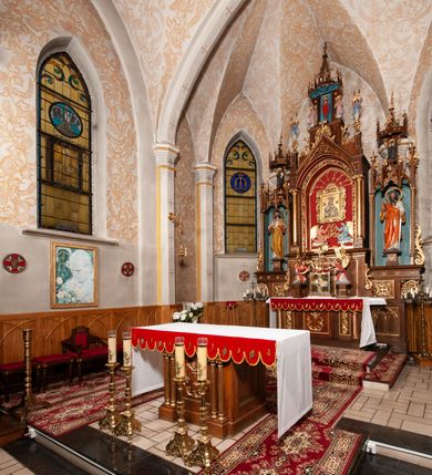 Zdjęcie nr 24: Ołtarz jednokondygnacyjny na wysokim, schodkowym cokole, trójosiowy, z wysuniętą mensą ołtarzową. Płyciny cokołu zdobione dekoracją w formie ostrołukowych maswerków i rozet w kwadratowej ramie. Antepedium flankowane parą kolumn ustawionych na wysokich cokołach i kanelowanych, skręconych trzonach z kompozytowymi kapitelami. W polu dwie prostokątne płyciny i kwadratowa pośrodku ze stylizowaną wicią roślinną i małą uskrzydloną główką aniołka. Tabernakulum proste, w kształcie prostopadłościennej skrzynki, o złotych drzwiczkach z wytłaczaną formą równoramiennego krzyża i wypukłą srebrną rybą oraz koszem z chlebem. Po obu stronach, na niewysokich postumentach dwa klęczące anioły z wysokimi skrzydłami, w biało różowych szatach z fioletowym podbiciem (skrzydła stylizowane na husarskie). Anioł po lewej z kadzielnicą, a anioł po prawej z kielichem.
Nastawa ołtarzowa o trzech oddzielnych niszach, nakrytych gotycyzującymi baldachimami z pinaklami, ozdobionymi żabkami. Oś środkowa w formie niszy zamkniętej ostrołukowo z wimpergą wspartą na smukłych, złoconych kolumienkach o kapitelach kompozytowych. Wimperga ze złoconymi, profilowanymi płycinami, zwieńczona rzeźbionymi liśćmi akantu i wolutami. Na przedłużeniu kolumn postumenty z figurami aniołów w biało niebieskich szatach, ze złotymi, wydłużonymi skrzydłami. Na osi na postumencie, pod gotycyzującym baldachimem figura świętego biskupa w czerwonej szacie z krzyżem w dłoni pomiędzy dwoma aniołami w białych i różowych szatach. W niszy, na czerwonym tle obraz Matki Boskiej Nieustającej Pomocy w prostej ramie z dekoracją w formie wici roślinnej. Poniżej dwa płaskorzeźbione anioły trzymające ażurowy monogram maryjny. Anioły o pozłacanych skrzydłach, na chmurach, w różowej i niebieskiej szacie. Osie boczne w formie aediculi z gotycyzującym baldachimem. W niszach figury, po lewej św. Piotra, a po prawej św. Pawła. Aedicule ujęte są smukłymi, skręconymi kolumnami z kapitelami wolutowymi oraz roślinnym uszakiem z perełkowaniem. Nad nimi imposty, na których rzeźby osłaniane baldachimami, po lewej świętego w habicie jezuity, a po prawej św. Jana Kantego. Drewno bejcowane na ciemny brąz, z nakładanymi elementami dekoracyjnymi złoconymi. 