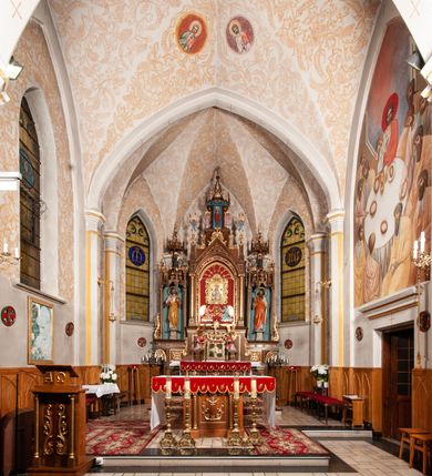 Zdjęcie nr 34: Ołtarz jednokondygnacyjny na wysokim, schodkowym cokole, trójosiowy, z wysuniętą mensą ołtarzową. Płyciny cokołu zdobione dekoracją w formie ostrołukowych maswerków i rozet w kwadratowej ramie. Antepedium flankowane parą kolumn ustawionych na wysokich cokołach i kanelowanych, skręconych trzonach z kompozytowymi kapitelami. W polu dwie prostokątne płyciny i kwadratowa pośrodku ze stylizowaną wicią roślinną i małą uskrzydloną główką aniołka. Tabernakulum proste, w kształcie prostopadłościennej skrzynki, o złotych drzwiczkach z wytłaczaną formą równoramiennego krzyża i wypukłą srebrną rybą oraz koszem z chlebem. Po obu stronach, na niewysokich postumentach dwa klęczące anioły z wysokimi skrzydłami, w biało różowych szatach z fioletowym podbiciem (skrzydła stylizowane na husarskie). Anioł po lewej z kadzielnicą, a anioł po prawej z kielichem.
Nastawa ołtarzowa o trzech oddzielnych niszach, nakrytych gotycyzującymi baldachimami z pinaklami, ozdobionymi żabkami. Oś środkowa w formie niszy zamkniętej ostrołukowo z wimpergą wspartą na smukłych, złoconych kolumienkach o kapitelach kompozytowych. Wimperga ze złoconymi, profilowanymi płycinami, zwieńczona rzeźbionymi liśćmi akantu i wolutami. Na przedłużeniu kolumn postumenty z figurami aniołów w biało niebieskich szatach, ze złotymi, wydłużonymi skrzydłami. Na osi na postumencie, pod gotycyzującym baldachimem figura świętego biskupa w czerwonej szacie z krzyżem w dłoni pomiędzy dwoma aniołami w białych i różowych szatach. W niszy, na czerwonym tle obraz Matki Boskiej Nieustającej Pomocy w prostej ramie z dekoracją w formie wici roślinnej. Poniżej dwa płaskorzeźbione anioły trzymające ażurowy monogram maryjny. Anioły o pozłacanych skrzydłach, na chmurach, w różowej i niebieskiej szacie. Osie boczne w formie aediculi z gotycyzującym baldachimem. W niszach figury, po lewej św. Piotra, a po prawej św. Pawła. Aedicule ujęte są smukłymi, skręconymi kolumnami z kapitelami wolutowymi oraz roślinnym uszakiem z perełkowaniem. Nad nimi imposty, na których rzeźby osłaniane baldachimami, po lewej świętego w habicie jezuity, a po prawej św. Jana Kantego. Drewno bejcowane na ciemny brąz, z nakładanymi elementami dekoracyjnymi złoconymi. 